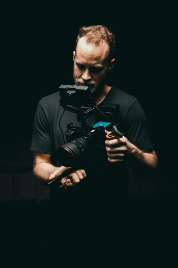 A man wearing black stands against a black background. He is operating a video camera.