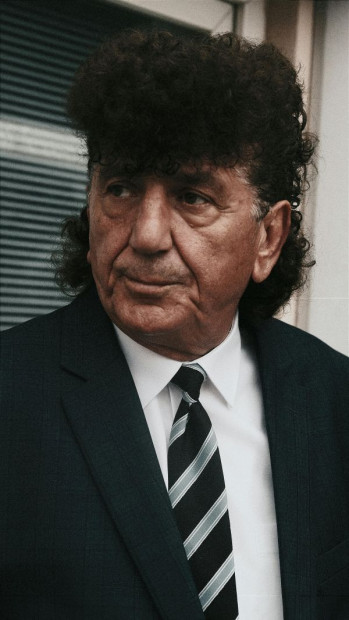 A man in a black business suit with a striped tie looks to the left. He has big black curly hair in a mullet.