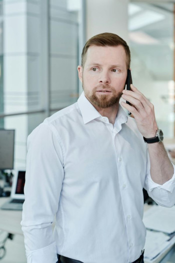 A white man with dark hair and a goatee stands talking on the phone in an office. He is wearing a white shirt.