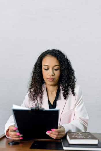 A black woman with long, dark, curly hair sits at a desk looking down at a file in her hand. She is wearing a black shirt and light coloured blazer.