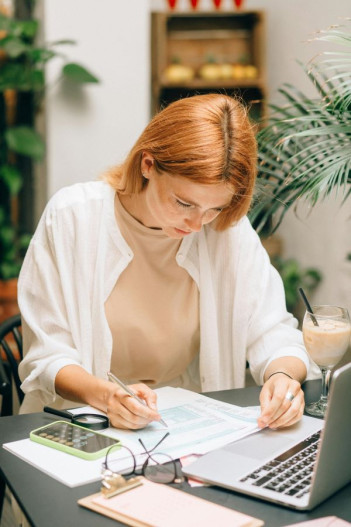 A red-headed woman sits looking down at papers on her desk. Her laptop sits in front of her.