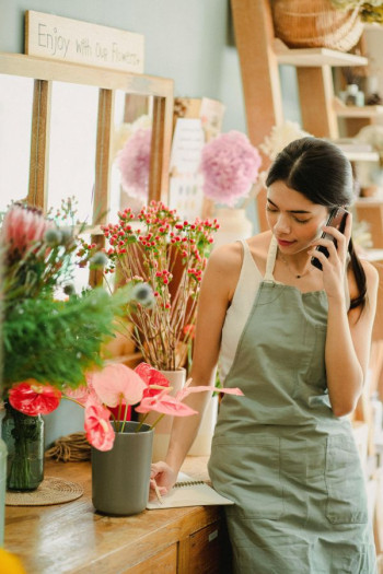 A woman with long dark hair in a ponytail leans against a desk in a florist. There are flowers around her. She is on the phone and writing down notes.