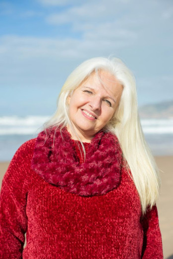 A middle aged woman with long platinum blonde hair stands on a beach. She is wearing a red jersey and red scarf.