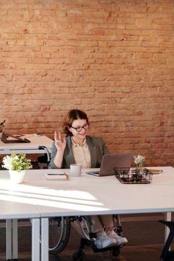 A red haired woman sits at a desk and waves at her laptop. She is in a wheelchair and has glasses.