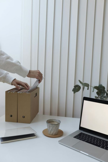 A person's arm in a white business shirt adds a file to a brown box. The box sits on a desk. Beside the box is a cup of coffee and an open laptop.