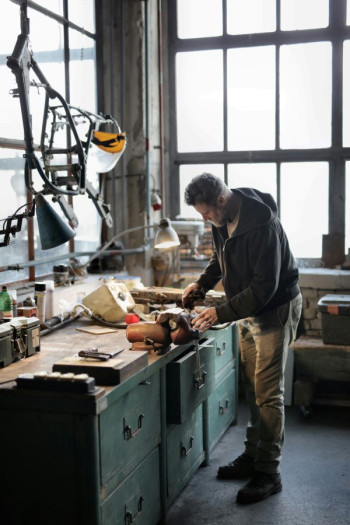 A man stands over a work desk in a garage. He has panelled windows behind him and in front of him. He is working on something in a vice. He wears boots, jeans, and a dark hoody. He has short brown hair.