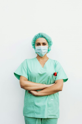 A female nurse stands looking at the camera with her arms folding. She is wearing green scrubs that includes a face mask and a hair net.