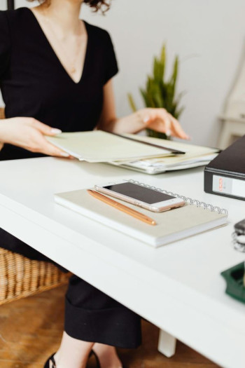 A woman wearing a v-neck black top sits at a desk. It has papers, notebooks, and a phone on it but looks organised. 