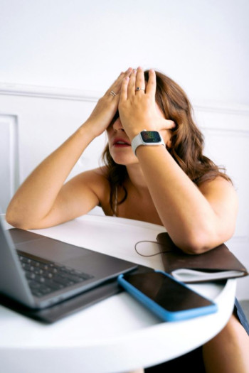 A woman sits at a desk in front of an open laptop with her head in her hands. She has an iPhone near her.