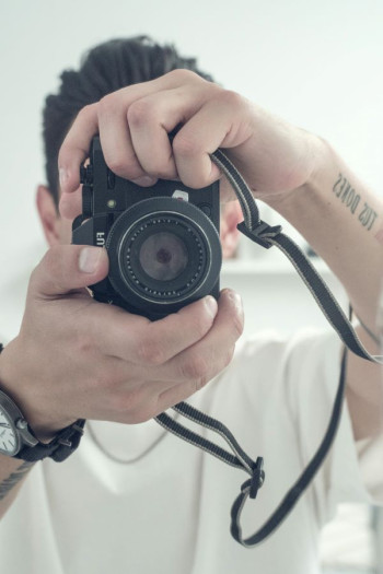 A man in a white t-shirt holds a camera up to his face. The camera is facing us.