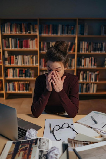 A woman with long dark hair tied up in a top knot sits at a desk in a lowly lit library. She wears a brown, long-sleeve top. She is rubbing her eyes. Her glasses are on the desk on top of papers. A laptop is open.