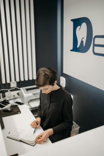 A person with short hair stands behind a receptionist desk looking at a diary. There is a dentist's logo on the wall behind them.