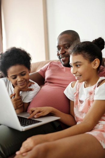 A black man in a red top watches a laptop. A boy is on this left and a girl is on his right and they are watching the laptop too.