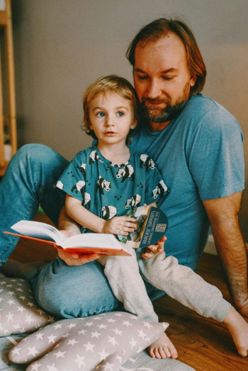 A father in a blue t-shirt and jeans sits reading a book to a child who is sitting on his knee