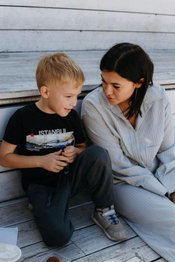A woman in a grey tracksuit and long, dark hair sits on a bench talking to a boy of about seven.