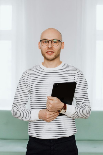 A bald man wearing a striped shirt and glasses stands holding an ipad