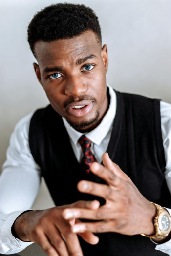A black man in a white shirt and black vest leans forward counting on his fingers