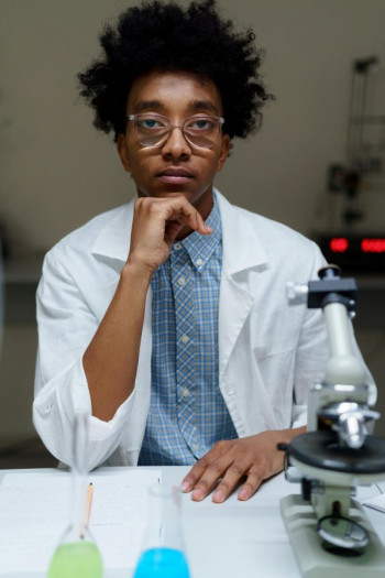 A black scientist looks at the camera. He is wearing clear reading glasses and a white lab coat.