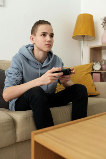 A teenage boy wearing a hoody and black jeans sits on the couch playing a games console.