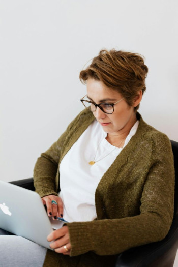 A woman with short hair and glasses sits with a laptop on her knee. She has an olive cardigan, jeans, and a white t-shirt on.