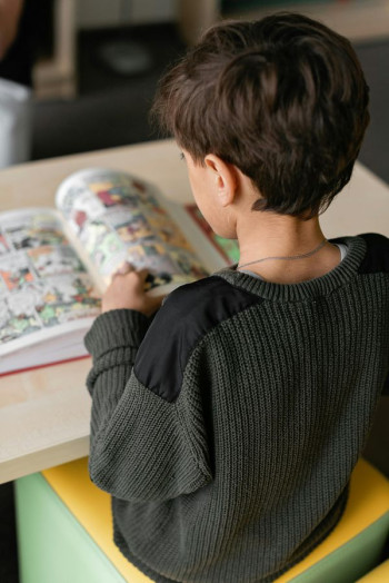 A small boy sits with his back to the camera reading a comic book that is sitting on a desk