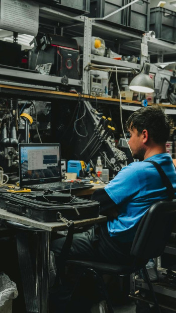 A repairman sits at a workstation that is surrounded by computer parts