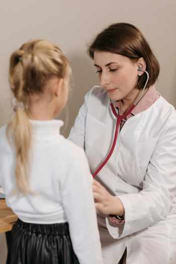 A doctor with a brown bob haircut uses a stethoscope to listen to a blonde child's chest