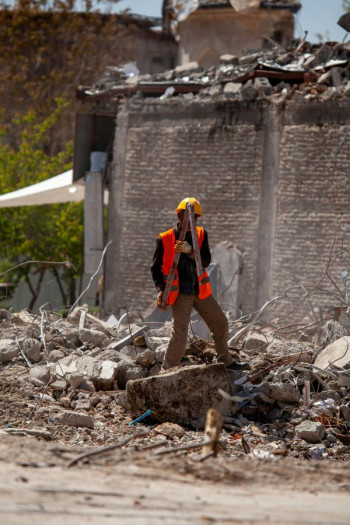 A man in a high vis vest with a yellow hard hat stands in the rubble of a building