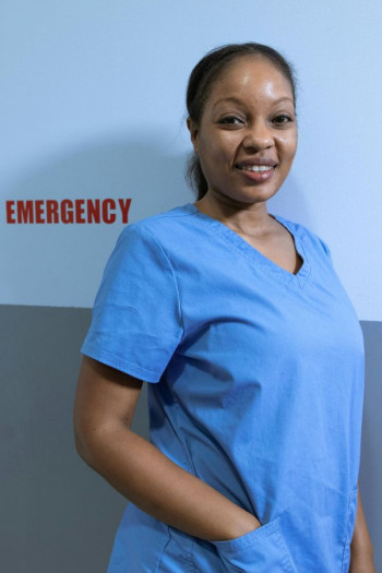 A black woman stands against a wall that says 'emergency.' She is wearing blue scrubs and has her hair tied back.