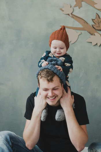 A father in a black t-shirt sits cross-legged on the floor. He has a small child on his shoulders.
