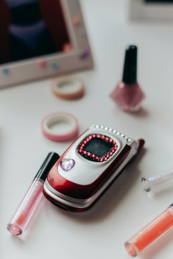 A flip phone sits on a table. It is bedazzled with some jewels and surrounded by lip gloss and stationery.