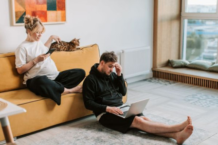 A white couple, a man and a woman, sit on the couch with a cat. They are wearing white t-shirts and black trousers. The man is on a laptop. 