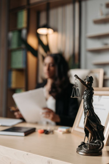 A blurred photo of a lawyer sitting at a desk. The focus is on a statue of the scales of justice on the right of the image.