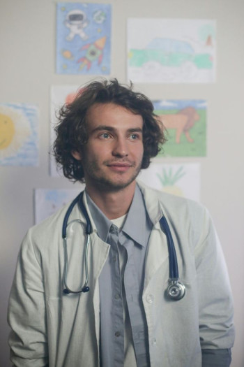 A white male doctor with dark shaggy hair stands near a wall. He has a white coat and stethoscope