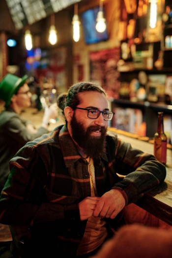 A man with dark hair in a bun sits at a bar. He is talking to someone unseen and dressed in casual clothing.