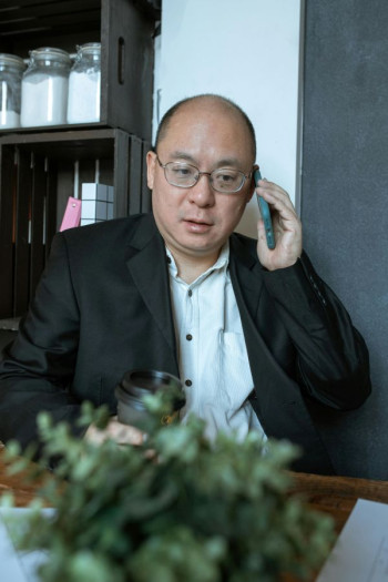 A bald Asian man with glasses sits speaking on a smartphone