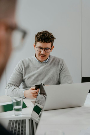 A white man with short dark curly hair and a moustache sits at a desk looking at his phone. He has glasses and a grey jersey. He is sitting behind a laptop on a desk and another person is opposite him.