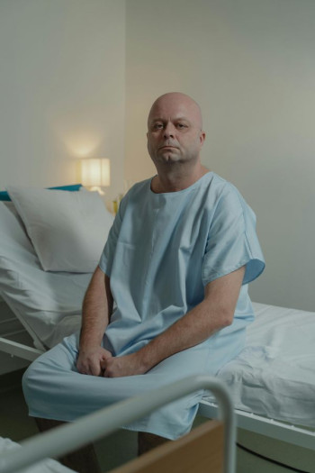 A bald white man sits on a bed in a hospital gown.