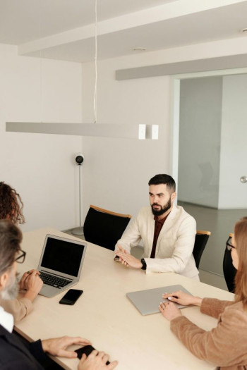 A white man with dark hair and beard wearing a light coloured suit sits at a table. He is being interviewed by three people