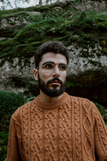 A man with dark hair and beard stands in a brown knitted jersey looking into the distance. He wears glasses and looks serious.