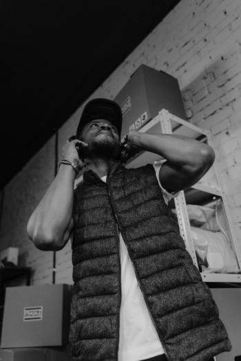 A man in a vest is on the phone in a warehouse. He has a cap on his head and is surrounded by boxes.