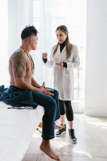 A shirtless man with tattoos sits on a medical bed talking to a doctor. She has long mousey-brown hair and wears a white coat.