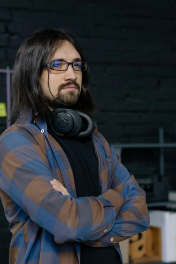 A man with long dark hair and a beard stands looking into the distance. He has glasses, headphones around his neck, and a checked blue and brown shirt.