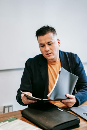 A man with short spikey dark hair sits at a desk holding a file. He has an orange t-shirt and dark blue blazer.