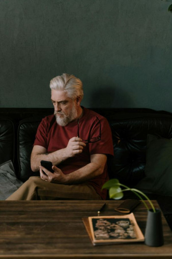 A man with silver, swept back short hair and beard sits on a couch behind a table. He is looking to the side and holding a smartphone. He is wearing a maroon t-shirt and beige trousers.