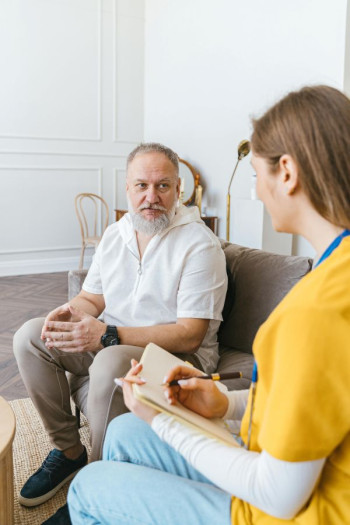 An older man with white hair and facial hair sits on a couch listening to a younger woman who is holding a clipboard.