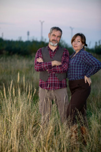 A middle aged couple stand in a wheat field wearing trousers and flannel checked shirts