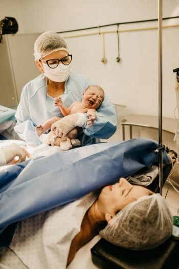 A woman lies down having delivered a baby by cesarean. A midwife holds the baby up to show the mother.  