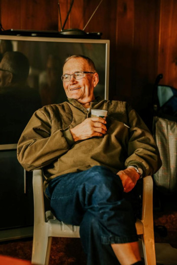 An older man with glasses sits in a plastic chair. He is relaxed, wearing a brown jersey, blue jeans, and glasses and holds a cup.