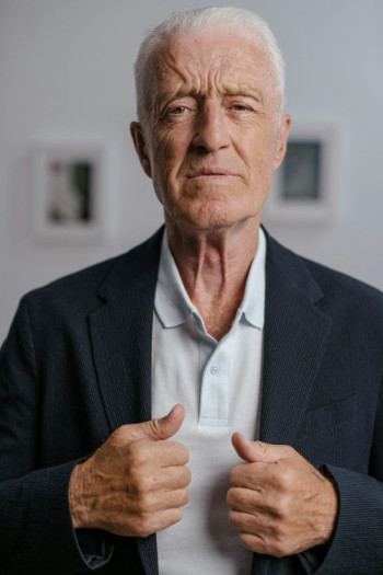 A white older man stands looking at the camera. He has white, short hair and a dark blazer with a polo neck tee.